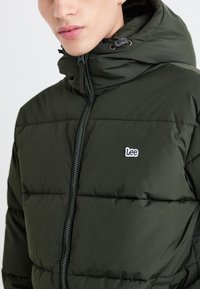 Giacca puffer verde scuro con zip frontale, dotata di cappuccio e piccolo logo bianco "Lee" sul petto. Texture liscia e design trapuntato.