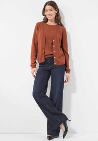 Vrouw in een roestkleurige cardigan en trui, met donkere wijde been jeans en zwarte puntige hakken, staand tegen een witte muur.