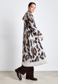 Cardigan lungo lavorato a maglia di colore bianco con stampa leopardata marrone e nera. Presenta un colletto a scialle e tasche laterali. Indossato sopra pantaloni neri e stivali.
