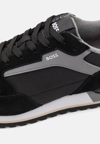 Zapatilla deportiva negra con una combinación de ante y tejido texturizado, con un panel lateral gris, diseño de cordones y detalle en la suela blanca.