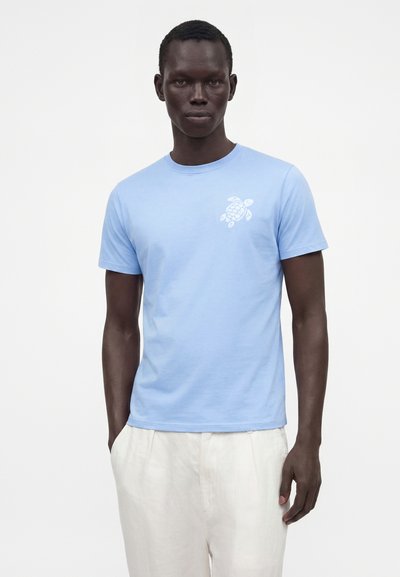 Homme portant un t-shirt bleu clair avec un motif de tortue blanche sur la poitrine et un pantalon écru, debout devant un fond uni.
