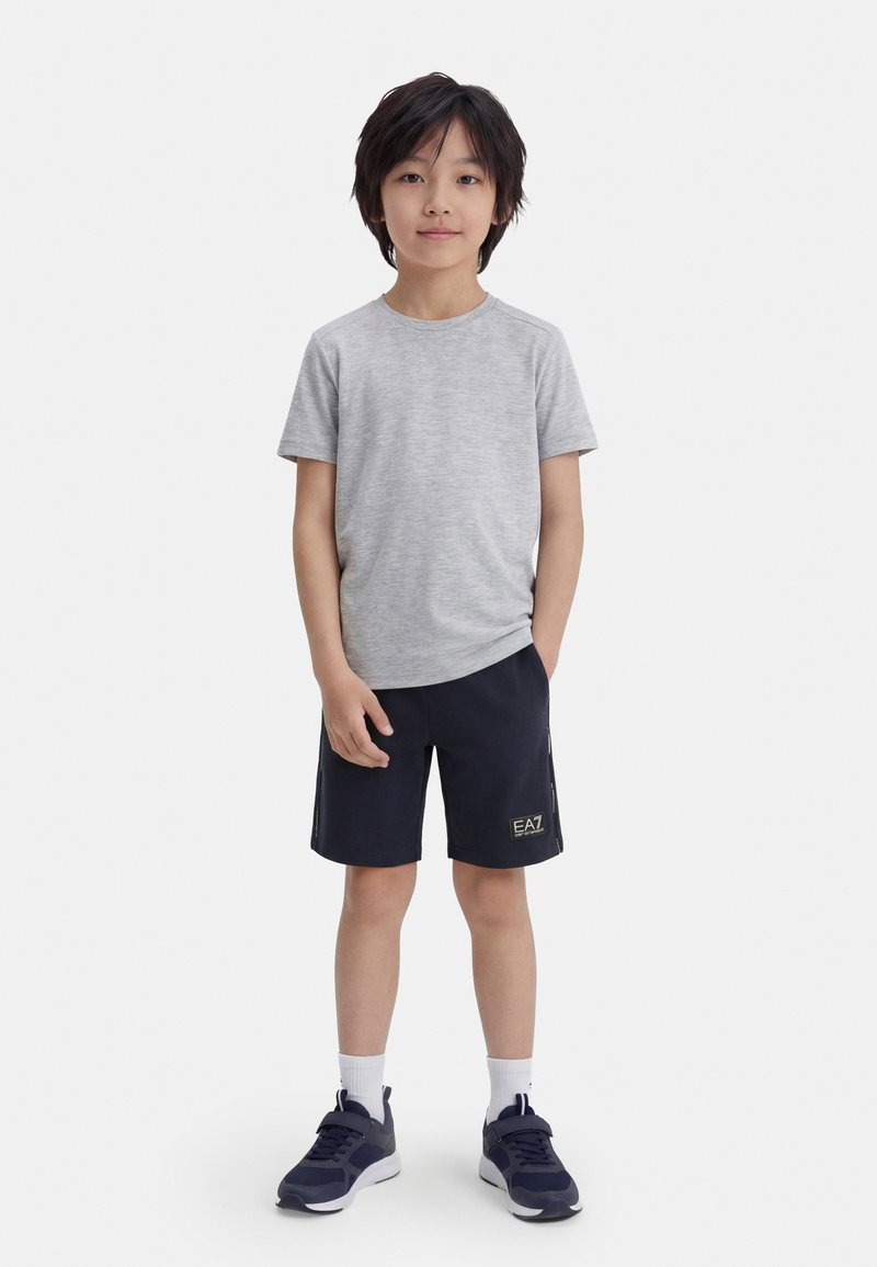 Το αγόρι στέκεται με το πρόσωπο προς τα εμπρός, φοράει ανοιχτό γκρι μπλουζάκι, σκούρα μπλε σορτς με λογότυπο, λευκές κάλτσες και μπλε αθλητικά παπούτσια σε λευκό φόντο.