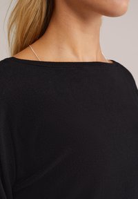 Haut en maille noire avec un décolleté bardot, tissu texturé et une subtile brillance. Un délicat collier en chaîne argentée est visible.