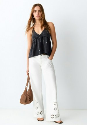 Mujer joven con top negro sin mangas y pantalones blancos de pierna ancha con recortes florales, sosteniendo un bolso marrón al hombro sobre fondo liso.