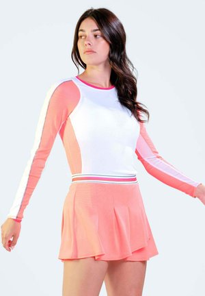 Frau trägt ein korallenfarbenes und weißes Langarm-Oberteil mit passendem korallenfarbenem, plissiertem Rock, schaut nach links mit leicht ausgestreckten Armen.