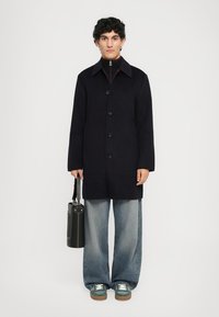 Navy wool coat, five buttons, relaxed fit. Layered collar with red accent, paired with light blue wide jeans and green sneakers.