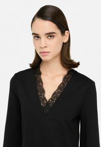 Zwarte top met lange mouwen en een diepe V-hals, voorzien van een delicate zwarte kantafwerking langs de halslijn. Soepele stoftextuur.