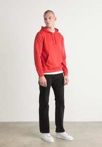 Felpa rossa in cotone con tasca a marsupio, strato interno bianco, abbinata a jeans neri e sneakers bianche, in piedi su uno sfondo neutro.