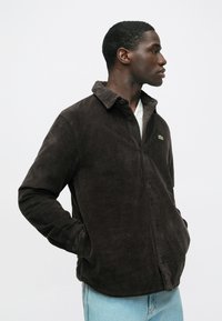 Sötétbarna kordbársony zakó gombos elülső résszel, hegyes gallérral és zöld logó díszítéssel, világoskék farmerrel párosítva.