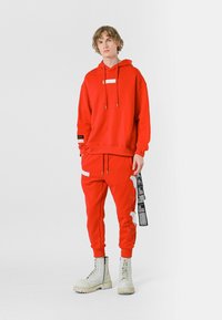 Sudadera roja con cordón y panel en contraste; pantalones a juego con acentos gráficos. Botas de combate blancas con suelas gruesas completan el conjunto.