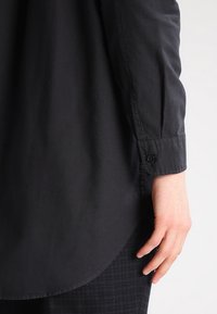 Camisa negra de manga larga hecha de una tela suave, con puños abotonados y un dobladillo redondeado, con una sutil textura y un ajuste entallado.