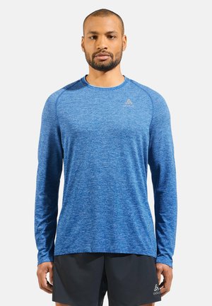 Langärmliges Sportshirt aus strukturiertem blauen Stoff, mit rundem Halsausschnitt und Logo-Akzent auf der Brust, kombiniert mit schwarzen Shorts.