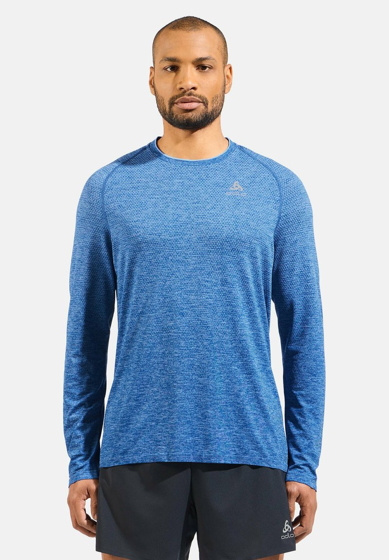 Langarm sportshirt van gestructureerde blauwe stof, met een ronde halslijn en een logo-accent op de borst, gecombineerd met zwarte shorts.