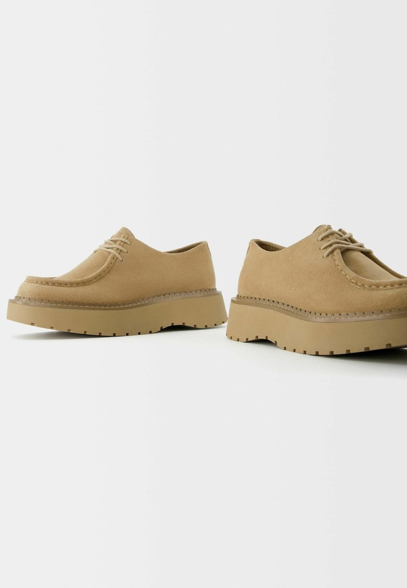Zapatos de ante beige con punta redondeada y cordones planos. Suelas gruesas con un patrón estriado que añaden tracción. Diseño simple y limpio sin adornos.