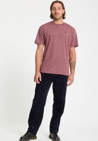 Camiseta de manga corta color marrón burdeos con cuello redondo y detalle de logo sutil, combinada con pantalones de pana en azul marino oscuro y zapatos negros.