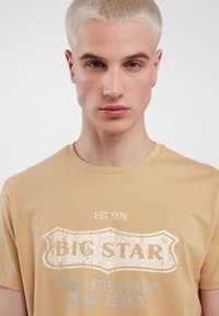 Smėlinis medvilninis marškinėliai su apvaliu iškirpimu. Priekyje yra nusidėvėjusi balta "BIG STAR" logotipas ir mėlynas tekstas "LEGENDINIAI MĖLYNI DŽINSAI".