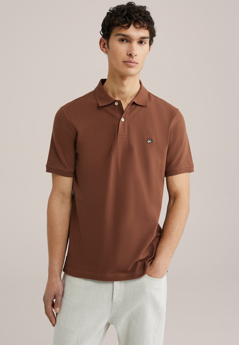 Brūna polo krekls ar gludu tekstūru, divu pogu slēgšana un neliels melns logs kreisajā krūtī. Saskaņots ar gaišiem biksēm.