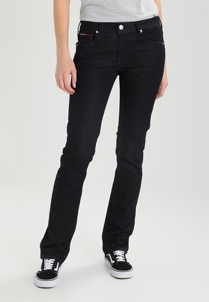 Personne portant un jean slim noir, un t-shirt gris et des baskets noires et blanches, debout devant un fond clair uni.