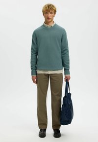 Jeune homme aux cheveux blonds bouclés portant un pull vert et un pantalon beige, tenant un sac fourre-tout bleu marine, se tenant devant un fond blanc.