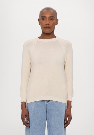 Maglione beige lavorato a maglia con scollo rotondo, polsini e orlo a coste. Presenta una finitura testurizzata e maniche raglan. Abbinato a jeans blu chiaro.