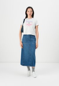 Šedé skrátené tričko s farebnou grafikou "Tommy Jeans", skombinované s džínsovou midi sukňou. Model má na sebe šedé ponožky a biele tenisky.
