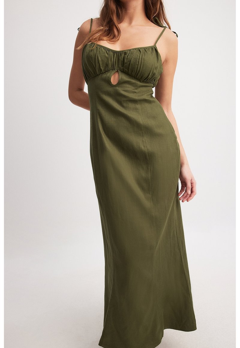 Femme portant une robe maxi sans manches vert olive avec une ouverture en forme de goutte sous la poitrine, debout devant un fond blanc uni.