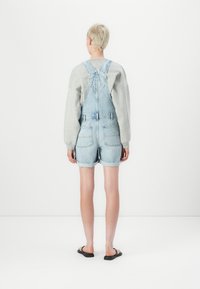 Salopette in denim azzurro chiaro, con bretelle regolabili, tasche anteriori e posteriori e orli arrotolati. Abbinata a una felpa grigia.