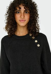 Zwarte gebreide trui met een ronde halslijn, voorzien van drie gouden knoopaccenten op één schouder en een zachte, gestructureerde afwerking.