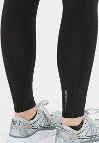 Schwarze Sportleggings, figurbetontes Design, glatte Textur, kombiniert mit grauen Laufschuhen mit orangefarbenen Akzenten und einem gemusterten Absatz.