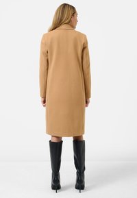 Camelkleurige jas met een rechte snit, gestructureerde stof en een gesneden kraag. Gecombineerd met hoge, zwarte laarzen met een stilettohak.