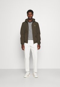 Giacca verde oliva con cappuccio, chiusura con zip e tasche frontali, abbinata a una maglietta a righe e pantaloni bianchi. Sneakers bianche completano il look.