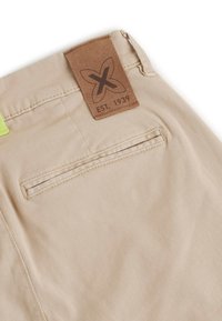 Pantalones cortos de tela beige con una etiqueta de cuero que presenta un logo y "EST. 1939", además de un bolsillo trasero con un zip oculto.
