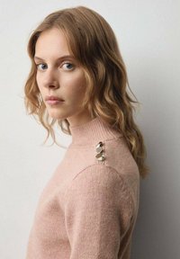 Svijetlo ružičasti pulover s visokim ovratnikom, izrađen od mekanog materijala; sadrži jedinstveni srebrni detalj na lijevoj strani ramena.