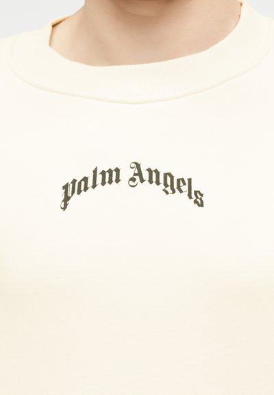 Λαιμός και άνω μέρος στήθους ατόμου που φοράει μπεζ φούτερ με μαύρο γοτθικό κείμενο «palm Angels» κεντραρισμένο κάτω από το γιακά.