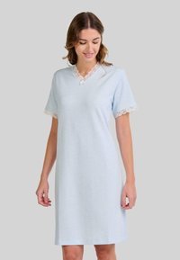 Mujer vestida con un pijama de rodilla azul claro, con detalles de encaje en las mangas y el escote, de pie frente a un fondo gris liso.
