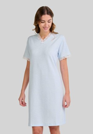 Mujer vestida con un pijama de rodilla azul claro, con detalles de encaje en las mangas y el escote, de pie frente a un fondo gris liso.