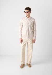 Camicia button-up a righe sottili in tonalità beige, abbinata a pantaloni crema e scarpe slip-on marroni. Accessoriata con occhiali da sole quadrati di grandi dimensioni.