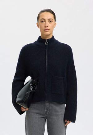 Femme aux cheveux tirés en arrière porte un pull zippé foncé et un jean gris, tenant un sac à main noir à son côté sur un fond uni.