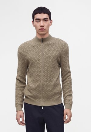 Jonge man met een lichtbruine gebreide trui met rits en donkere broek, staande tegen een effen witte achtergrond.