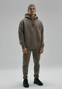 Zestaw bluzy z kapturem i spodni wykonany z przytłumionej brązowej tkaniny. Bluza ma kieszeń z przodu, czarny logo oraz regulowane sznurki. Czarne sneakersy.