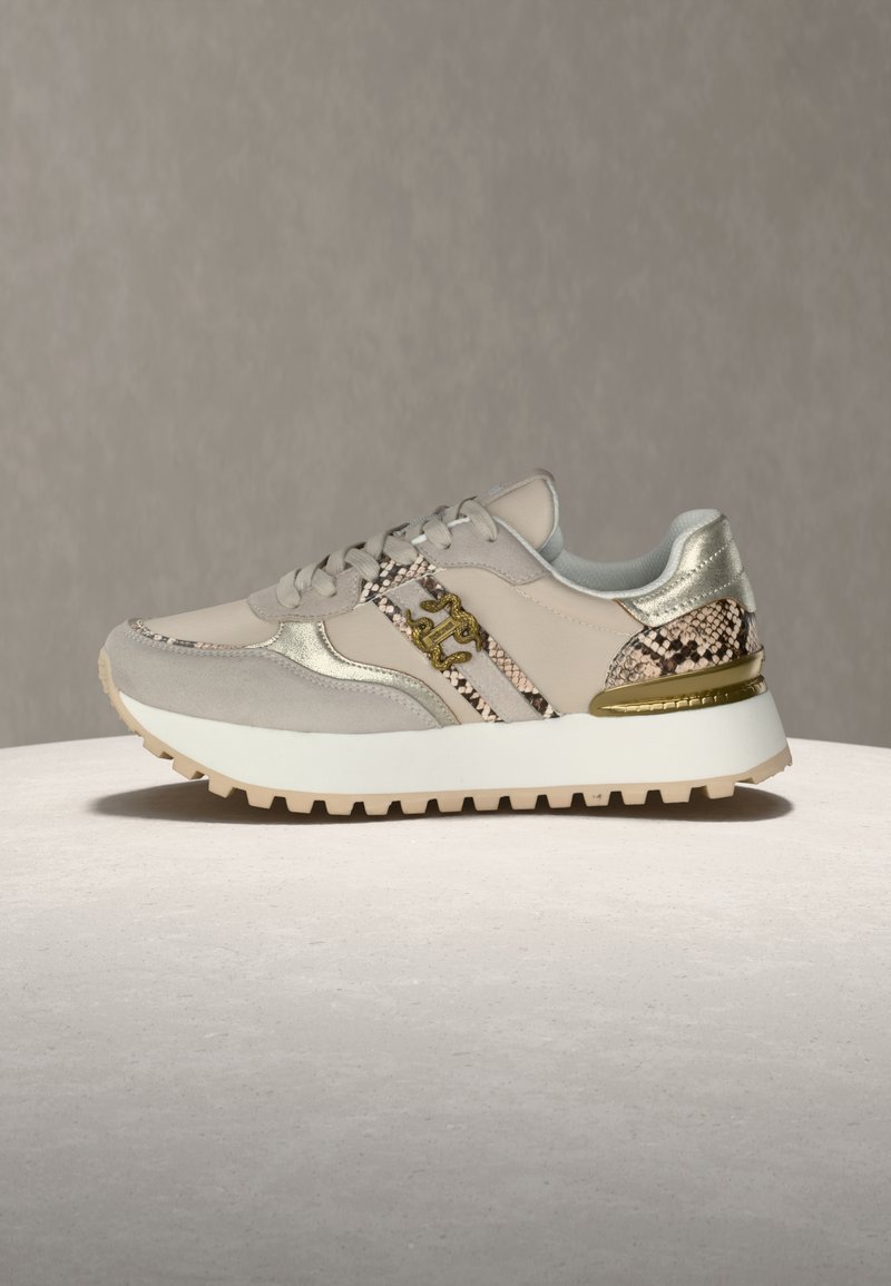 Μπέζ αθλητικό παπούτσι με σουέντ υλικό σε μπεζ αποχρώσεις και λεπτομέρειες με σχέδιο φιδιού, χρυσές διακοσμήσεις, χοντρή λευκή σόλα και μπεζ πέλμα, εμφανιζόμενο σε προφίλ πάνω σε ελαφρύ φόντο.