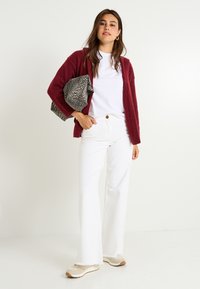 Gilet en peluche bordeaux sur un t-shirt blanc, associé à un jean évasé blanc et des baskets beiges. Tient un sac gris à motifs léopard.