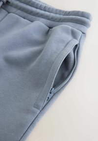 Gros plan sur une poche de pantalon de survêtement bleu avec une fermeture éclair partiellement ouverte et des coutures visibles sur un fond clair.