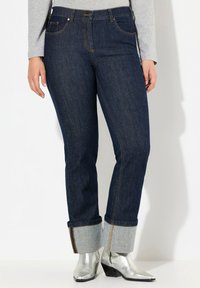 Jeans de mezclilla azul oscuro con costuras visibles, dobladillos enrollados que muestran tela gris y una cintura de altura media. Combinados con botas de tobillo plateadas.