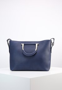 Námorně modrá kožená kabelka s zakřiveným vrchním úchytem se zlatými akcenty a odnímatelnými ramenními popruhy na bílém podstavci.