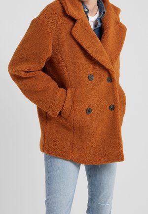 Personne portant un manteau teddy croisé couleur rouille avec poches latérales sur une chemise en denim et un jean bleu clair.