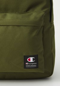 Olivinės spalvos audinio kuprinė su priekinėmis kišenėmis, ant kurios yra juodas logotipas su raudonu ir baltu emblemos tekstu: "Champion AUTHENTIC ATHLETICWEAR."