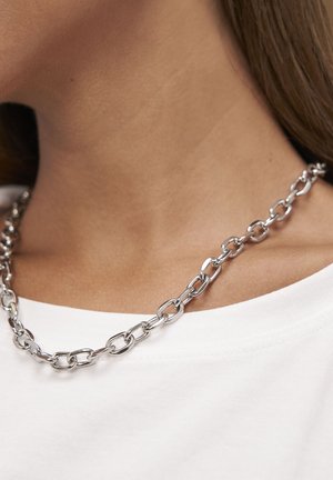 Sølvkæde med aflange led, højpoleret finish, båret over en hvid skjorte, hvilende på kravebensområdet.