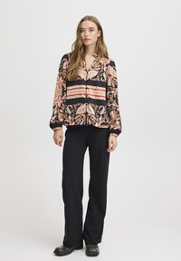 Blouse noire à manches longues avec motif floral et paisley dans des nuances de rouge, rose et or. Associé à un pantalon noir et des chaussures métalliques.
