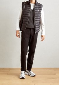 Hombre vestido con una camisa blanca de manga larga bajo un chaleco de punto gris y una chaqueta sin mangas acolchada negra, pantalones negros, zapatillas blancas y mochila negra.